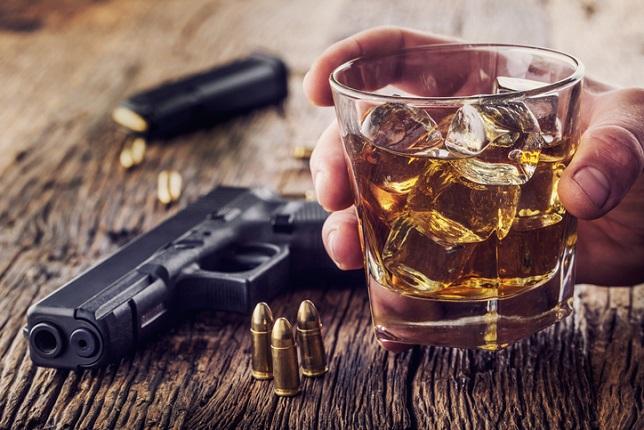 man's hand holding a drink, with a hand gun on a wooden table.