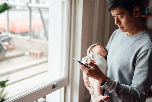 Man holding his baby, looking at his phone.