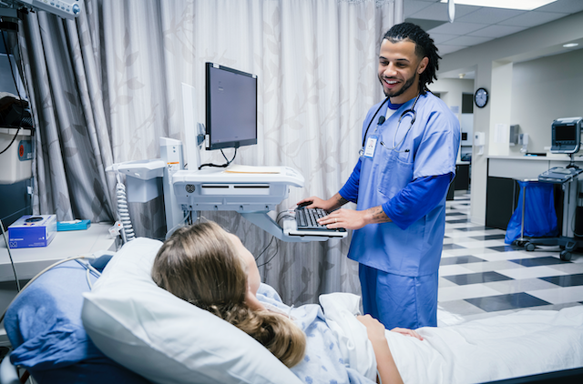 A nurse talks to a patient in a hospital bed while typing on a computer.