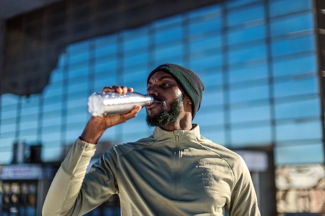 Man in hat drinks from aluminum water bottle while standing outdoors.