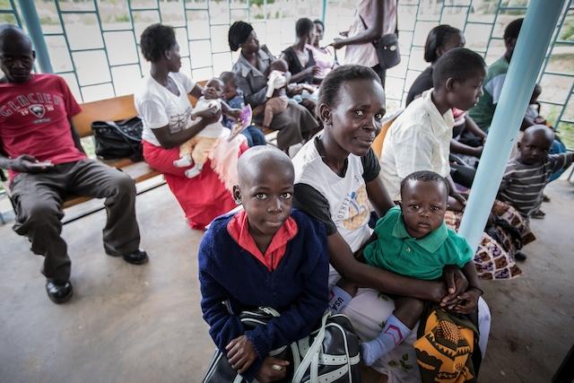 A mother sits with two young children in a crowded clinic waiting room.
