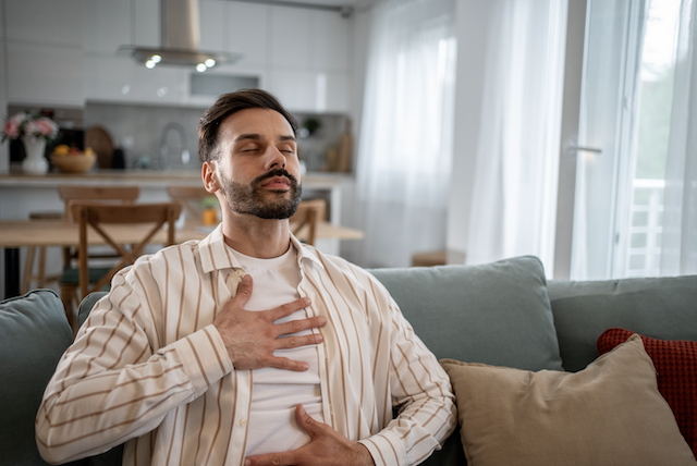 A man sits on a couch with his eyes closed, hands on his chest and abdomen, practicing diaphragmatic breathing.